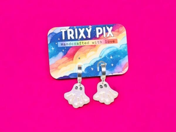 A pair of polymer clay ghost earrings attached to a colourful backing card against a bright pink background.