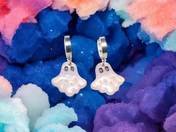A pair of polymer clay ghost earrings against a rainbow coloured cloudy background.