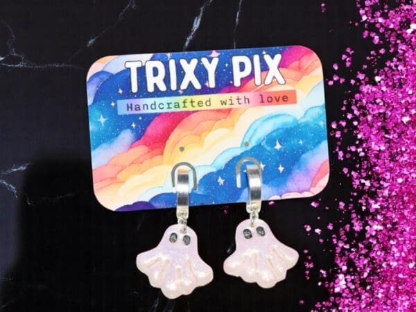 Polymer clay ghost earrings attached to a colourful backing card against a black background surrounded by pink glitter.
