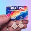 A female hand holds a pair of polymer clay ghost earrings attached to a colourful backing card.