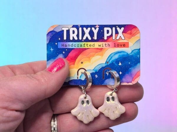 A female hand holds a pair of polymer clay ghost earrings attached to a colourful backing card.